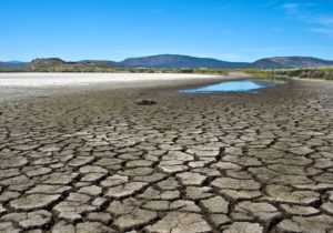 Cracked, dry and exposed lake bottom  in foreground of bed of Lower Klamath Lake, California with small patch of remaining water in background against backdrop of blue sky and distant barren hills.